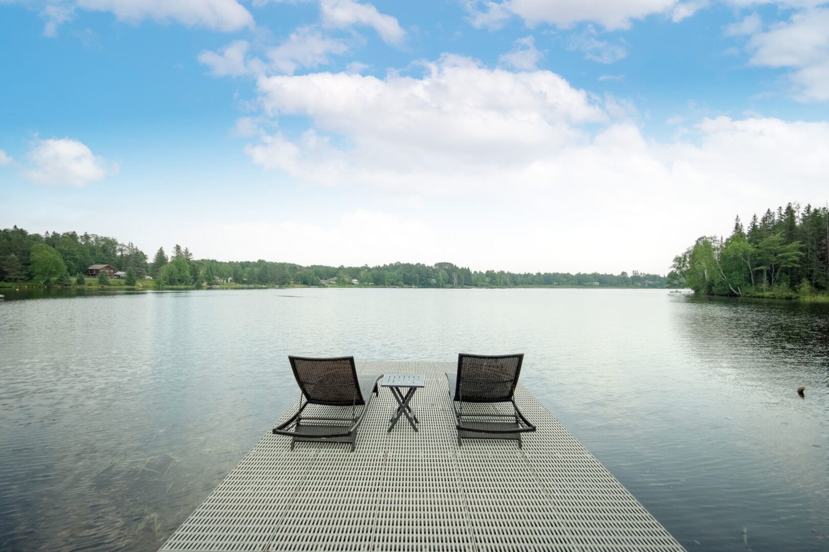 Two chairs sit on the end of a metal dock, looking out across a lake.