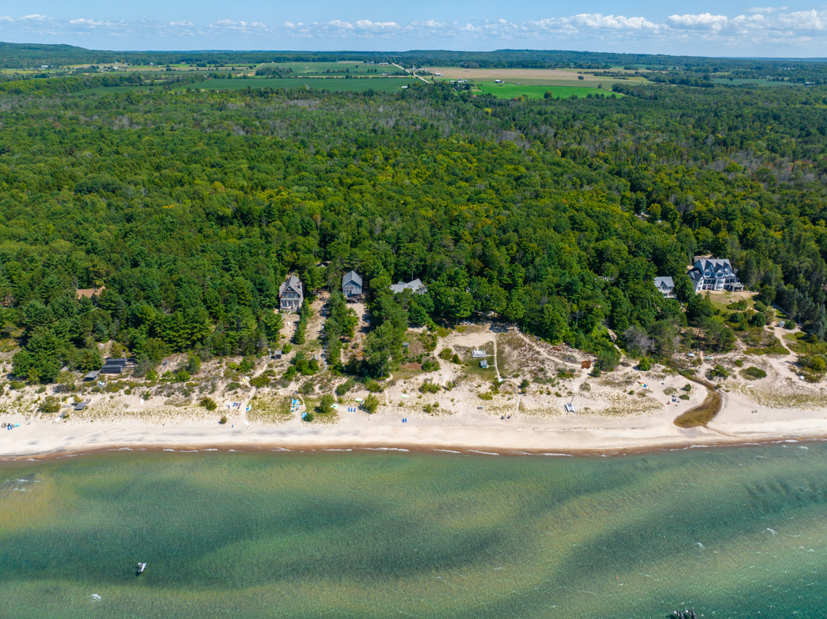 A shoreline with a sandy beach. Cottages sit up the shore, surrounded by green trees.