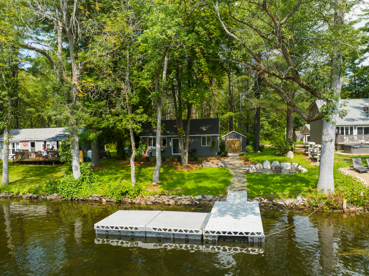 View from the water toward a small dock and a cottage on a grassy lot surrounded by green trees.