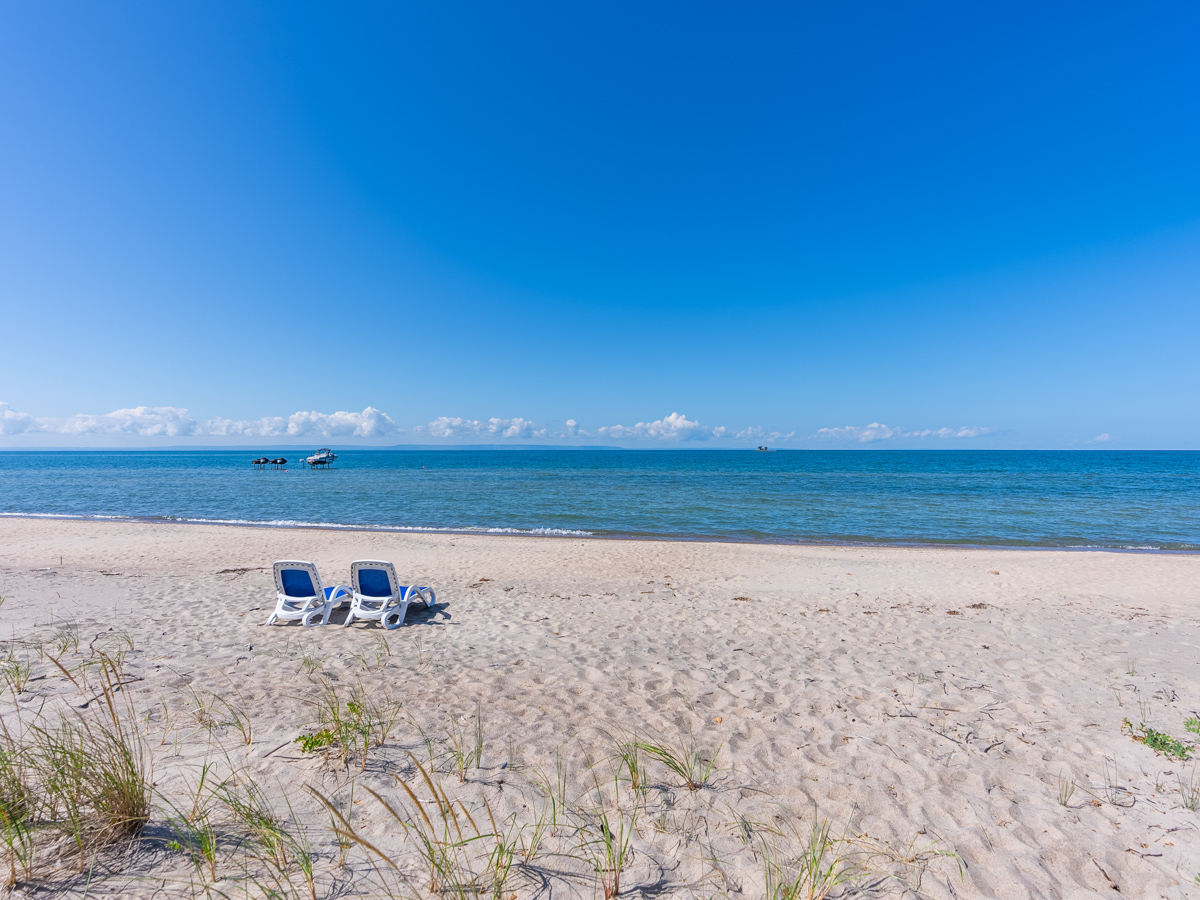 Two lounge chairs sit on a sandy beach in front of blue water.