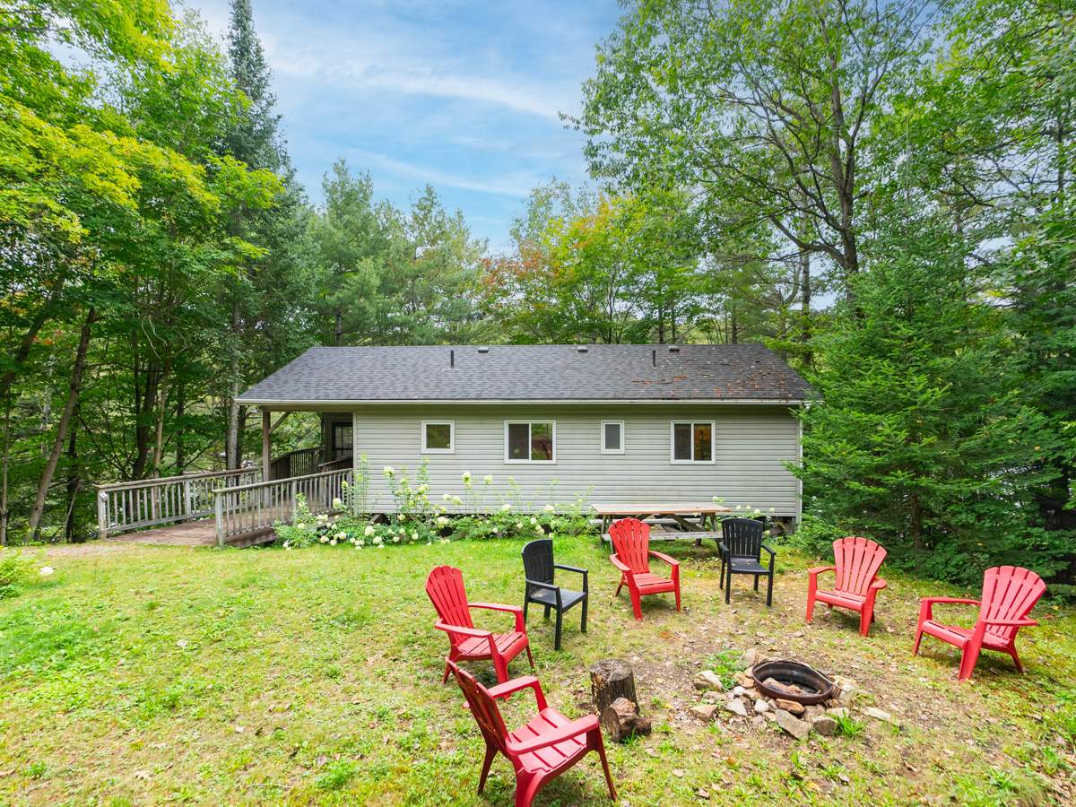 Red Muskoka chairs sit around a fire pit in front of a cottage surrounded by trees.