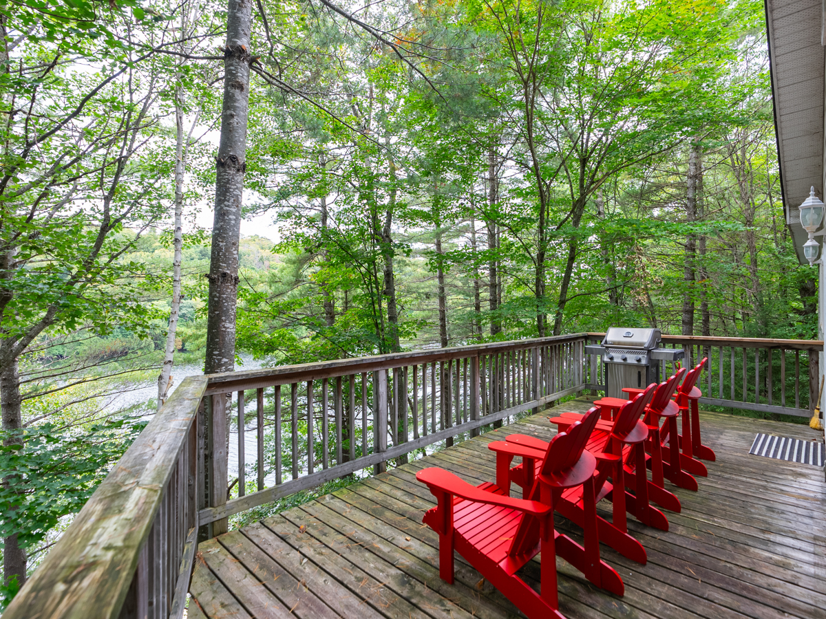 A wide deck with red Muskoka chairs looking out over a lake through green trees.