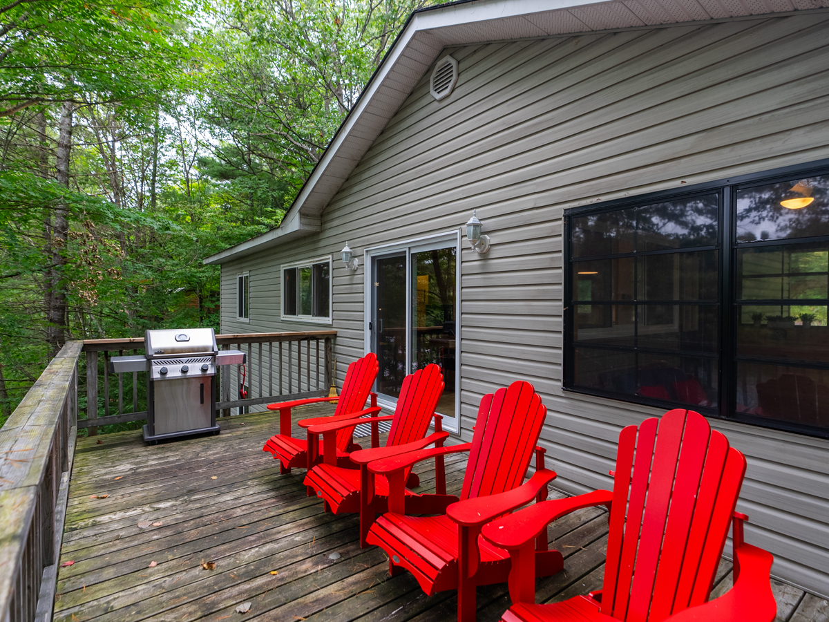 Four red Muskoka chairs and a barbecue sit on a deck off a cottage.
