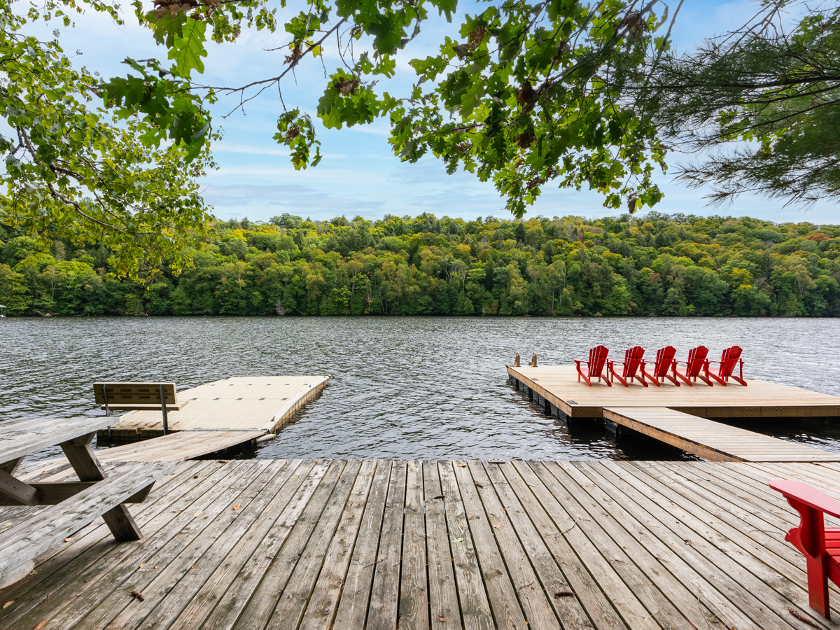 Two long pieces extend into a lake off a wide dock. Red Muskoka chairs sit at the end of one of the dock extensions, facing the water.