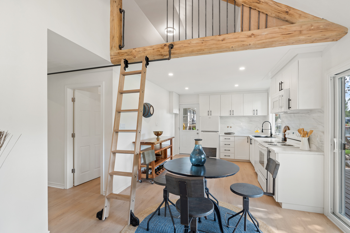 An open-concept cottage interior with dining an kitchen areas. A ladder leads up to a loft space above the kitchen.