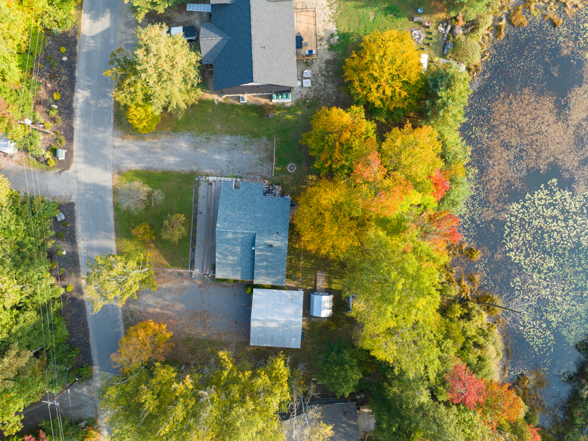 Aerial view of a bungalow cottage with a large driveway, surrounded by trees, in front of a small pond.