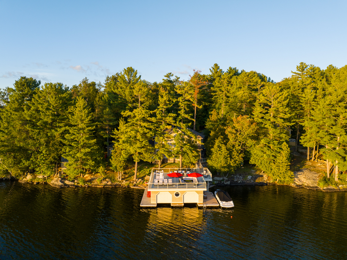 A large boathouse with a rooftop deck extends into a lake from a tree-lined shore.