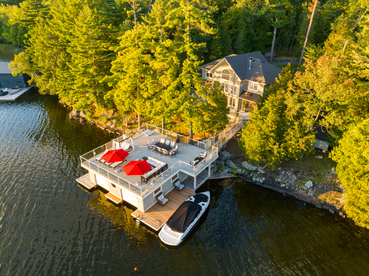 A large boathouse with a rooftop deck extends into a lake from a tree-lined shore.