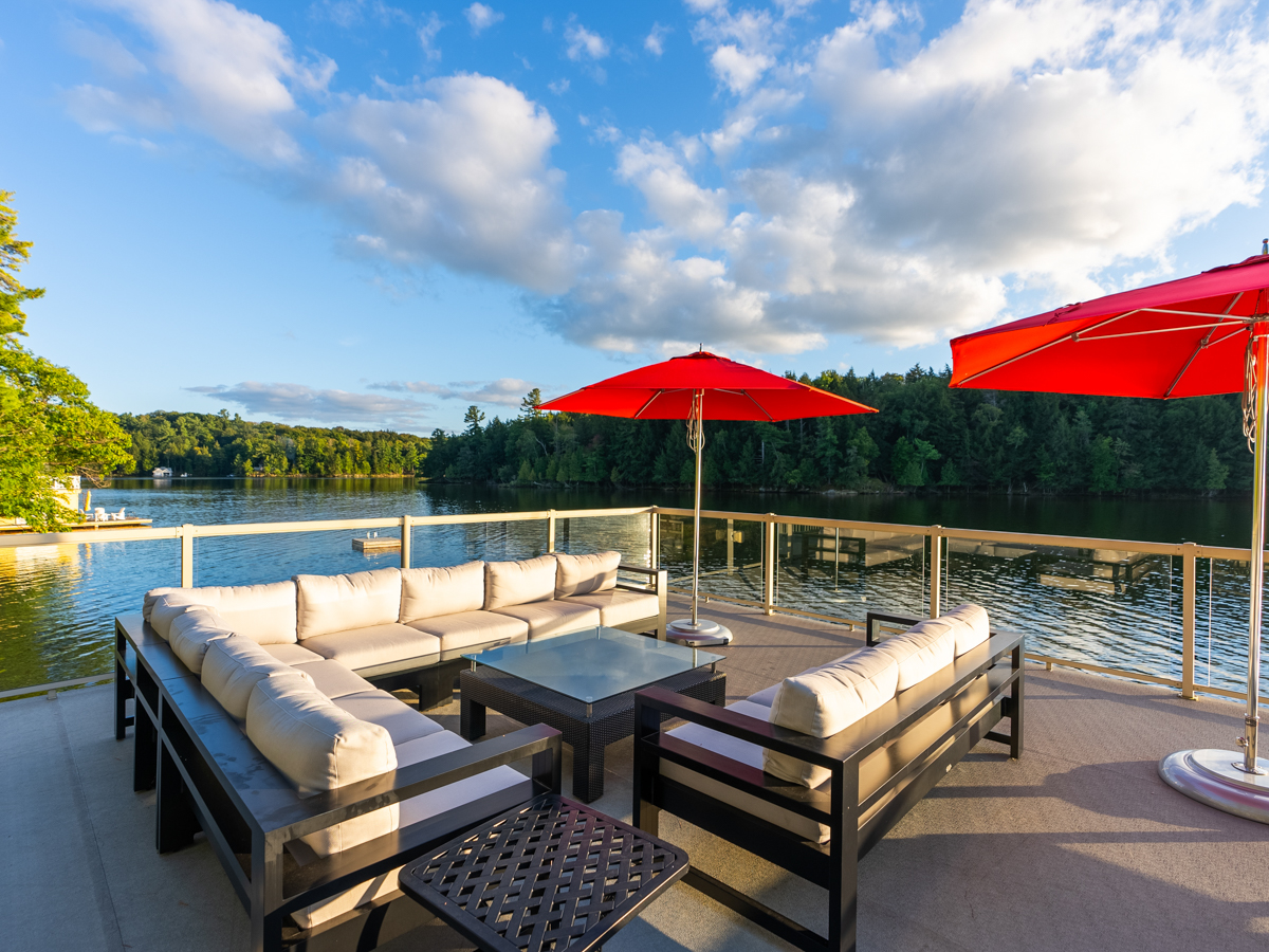 A large rooftop deck with outdoor living furniture and red sun umbrellas, looking out over a lake.