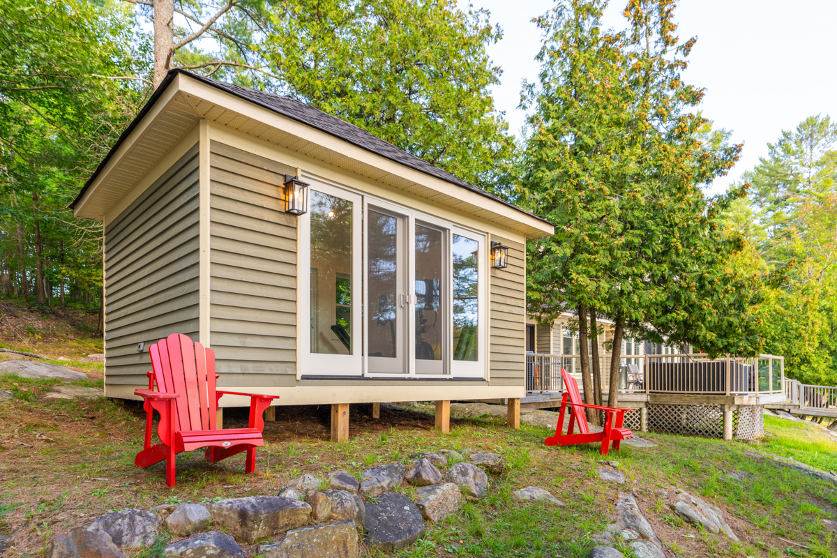 A small building with a large window and two red Muskoka chairs on either side, facing a lake.