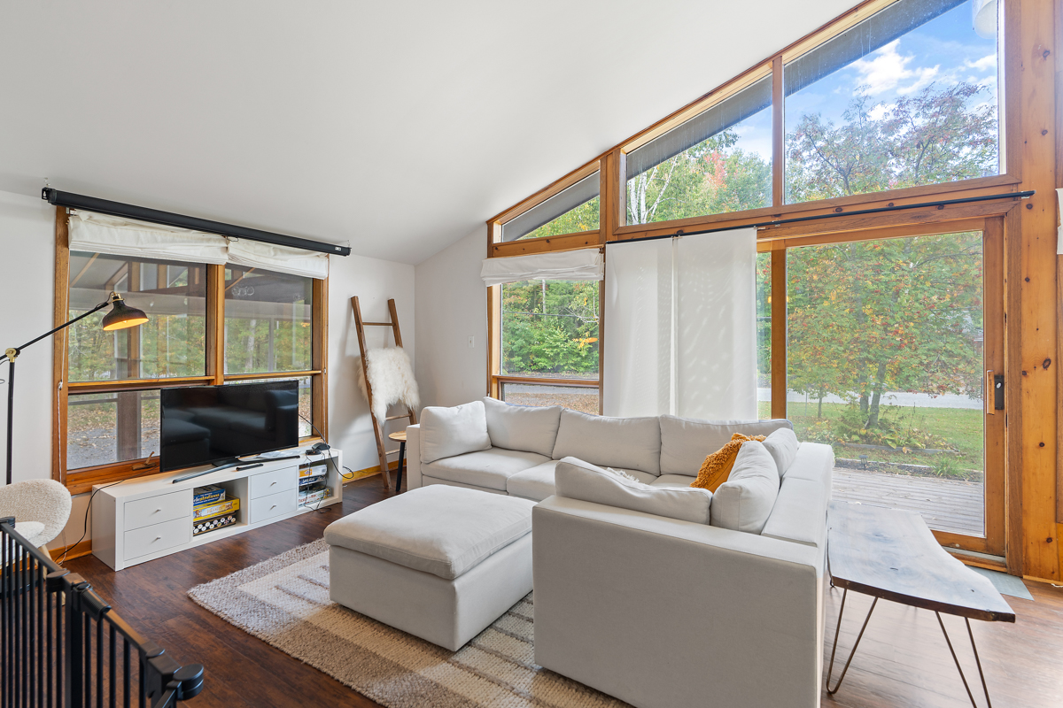 A bright living area with a white couch and a television. Floor-to-ceiling windows span the front wall of the house.