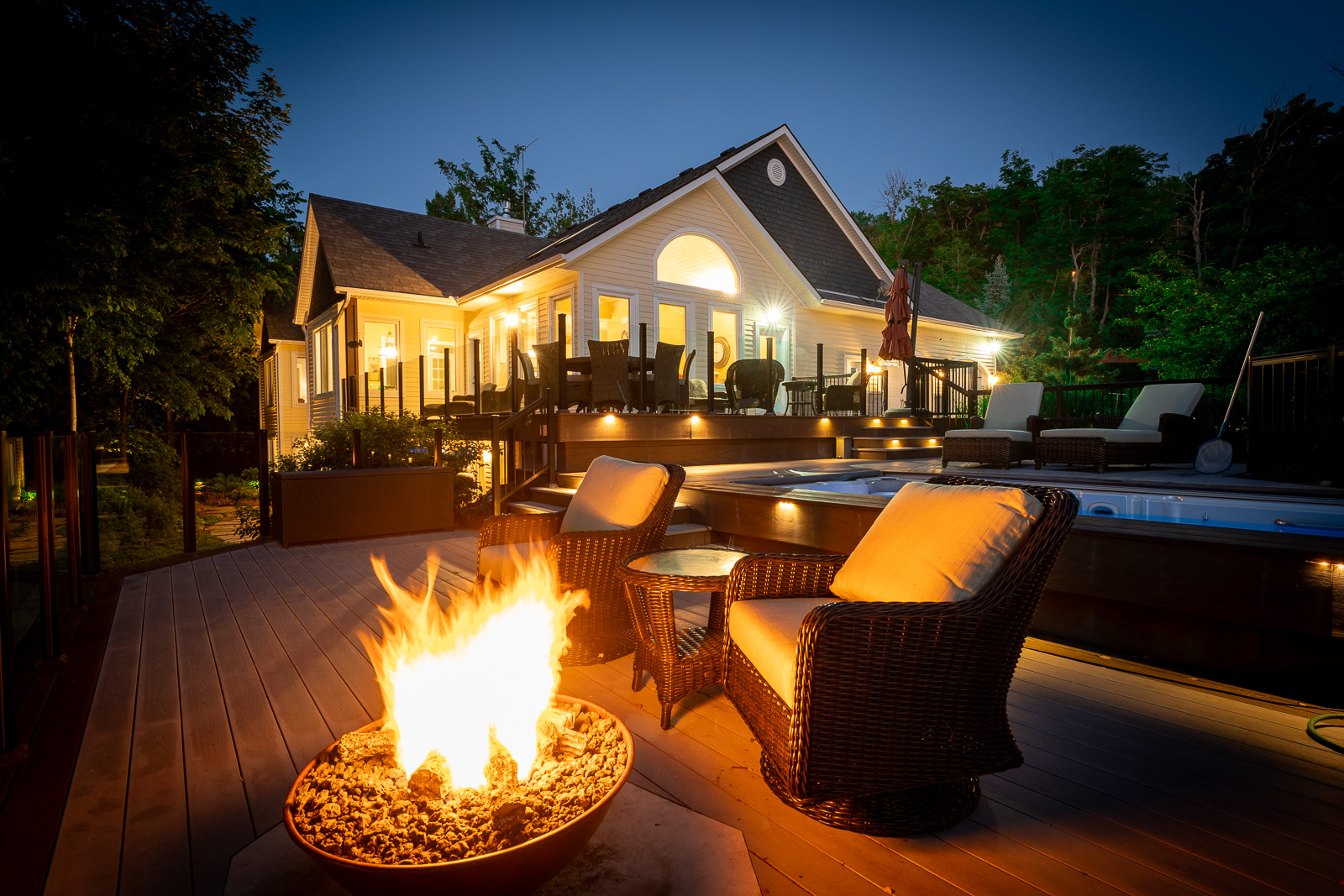 The deck of a large, luxury home at night, lit up with lights and a fire bowl.
