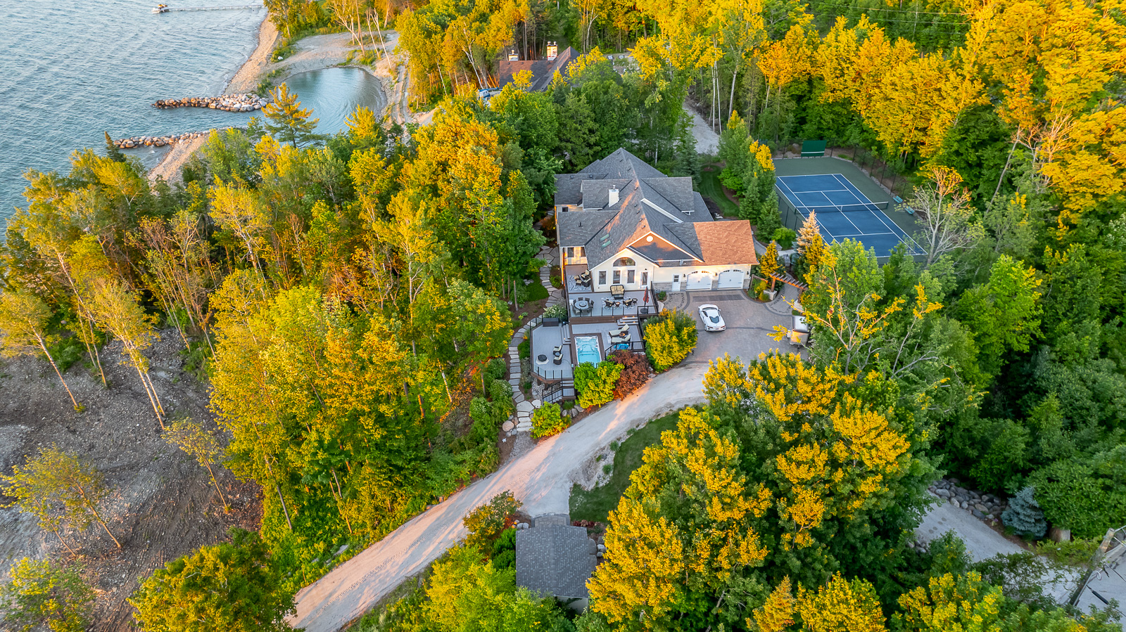 A large luxury property just off the waterfront with a big outdoor patio and a tennis court, surrounded by green trees.