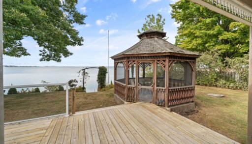 A gazebo sits on the edge of a large back deck of a cottage, with views of the lake.