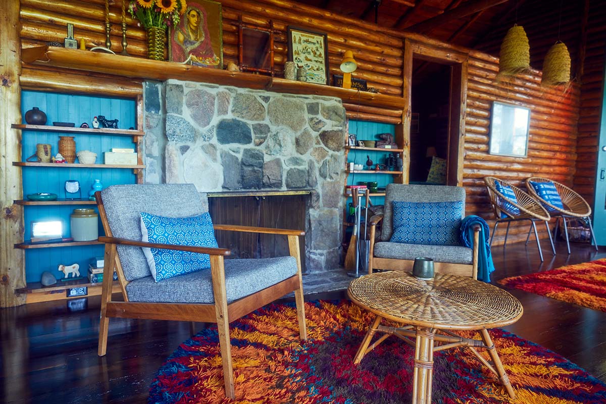 a photo of a log-walled cottage's living room with a stone fireplace, two chairs and a coffee table