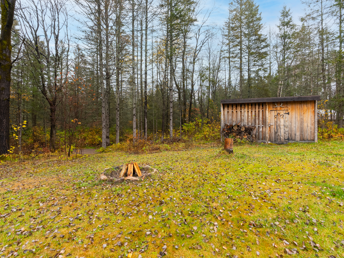 A small fire pit sits in a grassy yard, in front of a forested area. Behind it is a big wood shed.