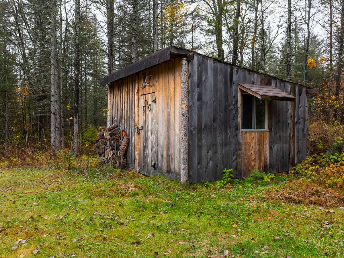 A big walk-in wood shed sitting in a grassy yard in front of a forested area.