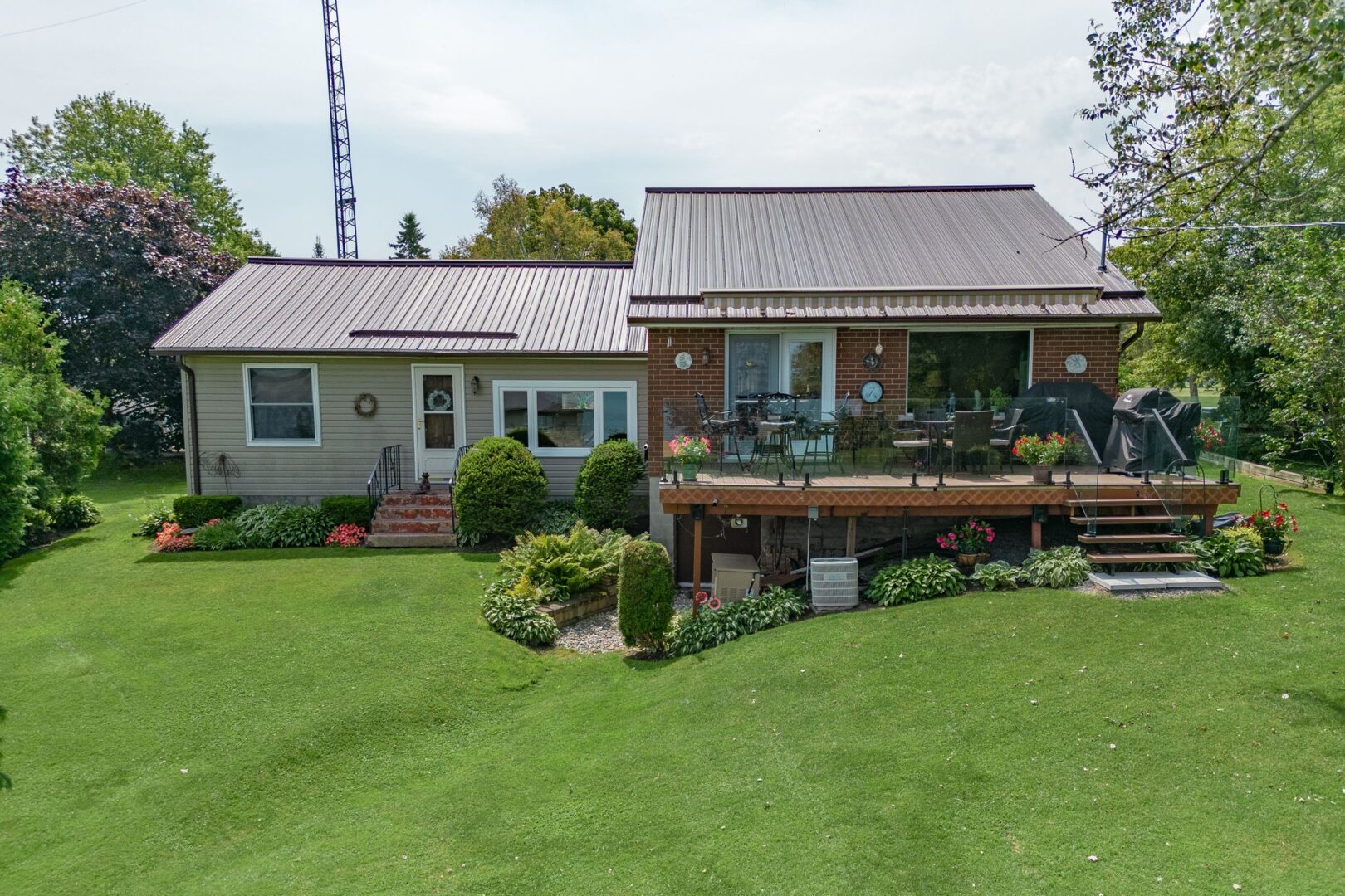 A large bungalow sits on a sloping grassy property. A deck with a glass railing extends off one side of the home.