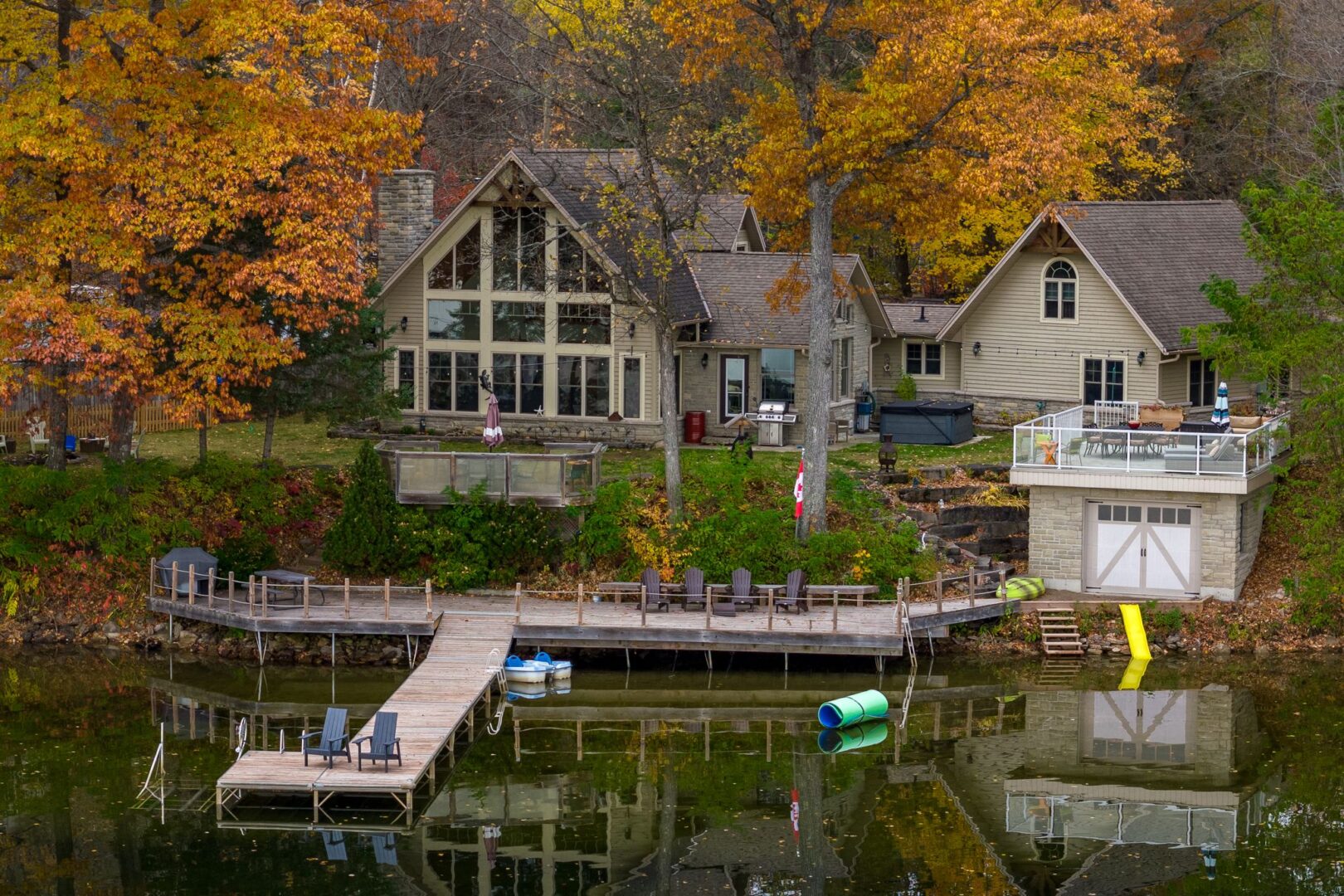 A large cottage with huge windows, a boathouse, and a long dock sits on the shore of a calm lake, surrounded by trees with colourful fall leaves.