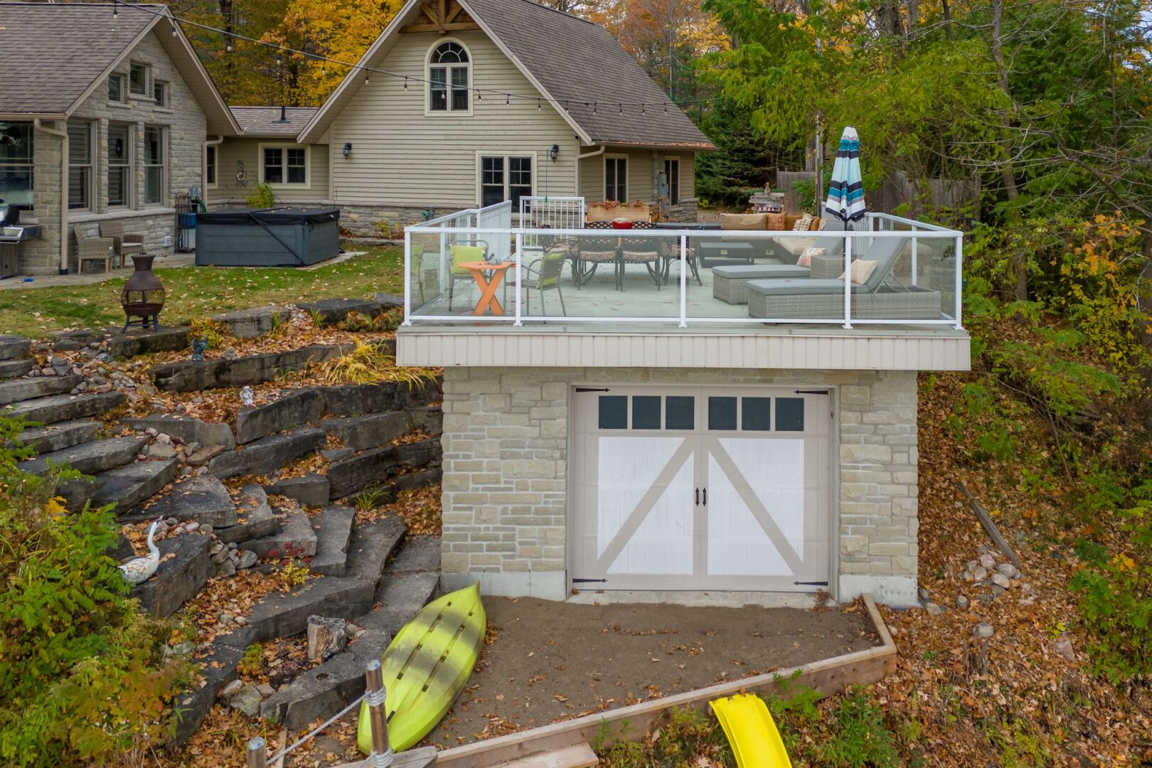 A large boathouse sits on the shore of a lake, in front of a large cottage. A big patio with outdoor furniture spans the roof of the boathouse.