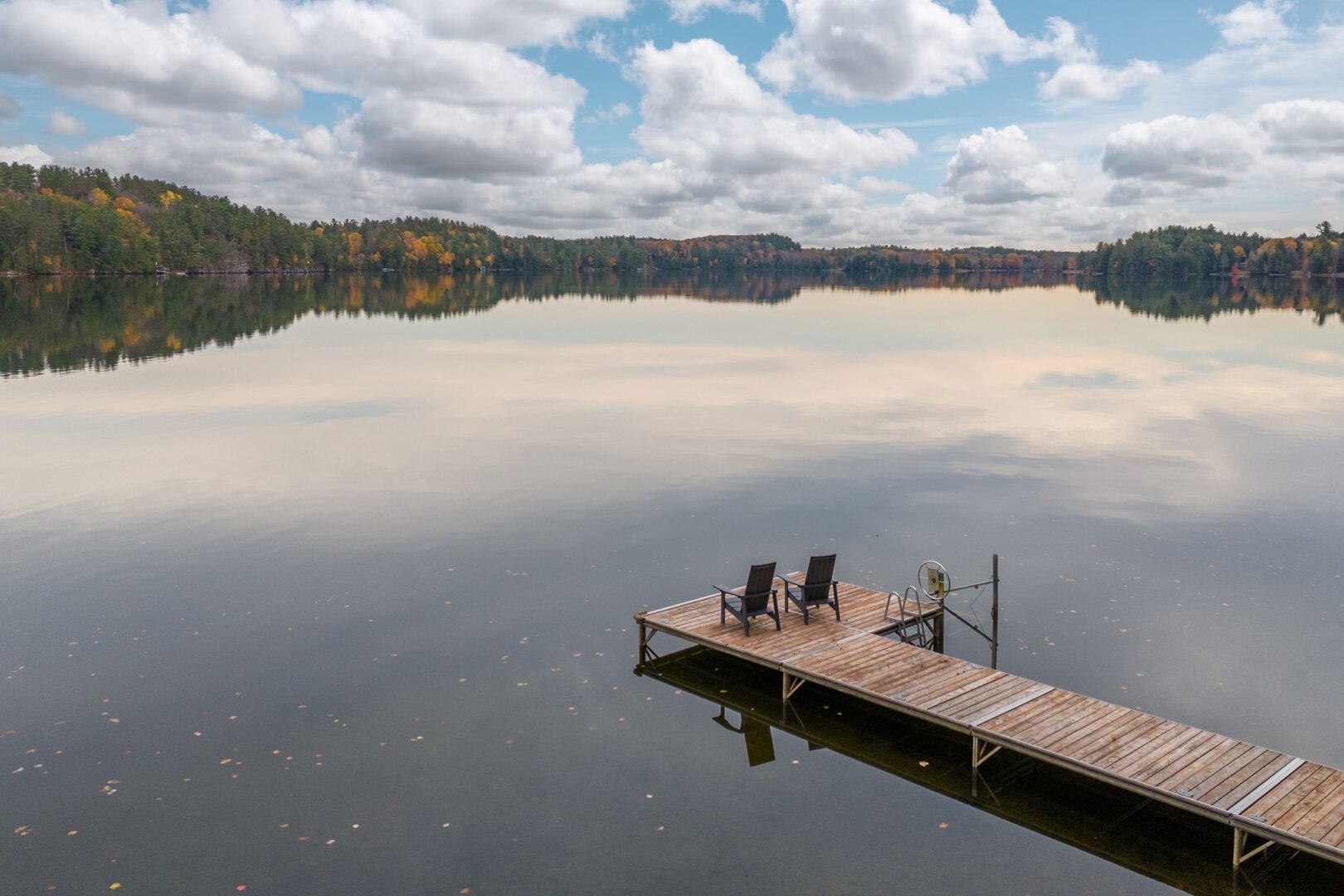 A long, narrow piece of dock extends out into the lake. Two Muskoka chairs sit on the end of the dock, facing the lake.