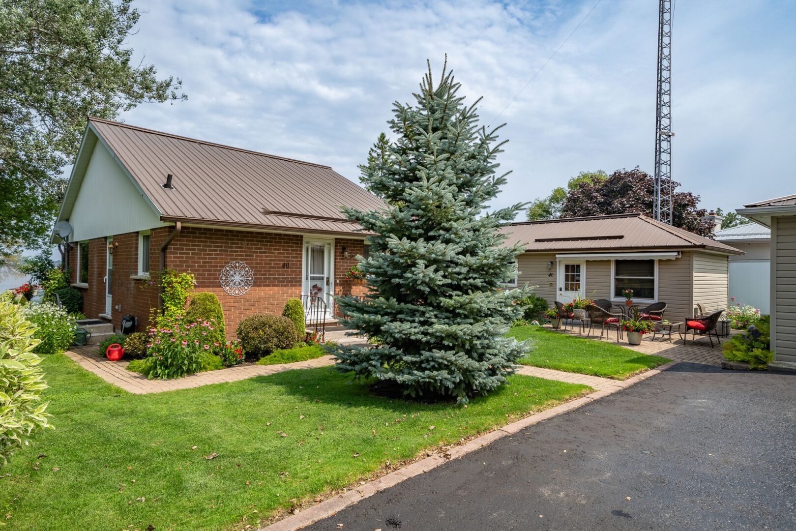 The front of a bungalow property on a rural, waterfront lot. A tall pine tree sits in the green space beside a paved driveway, in front of the home.