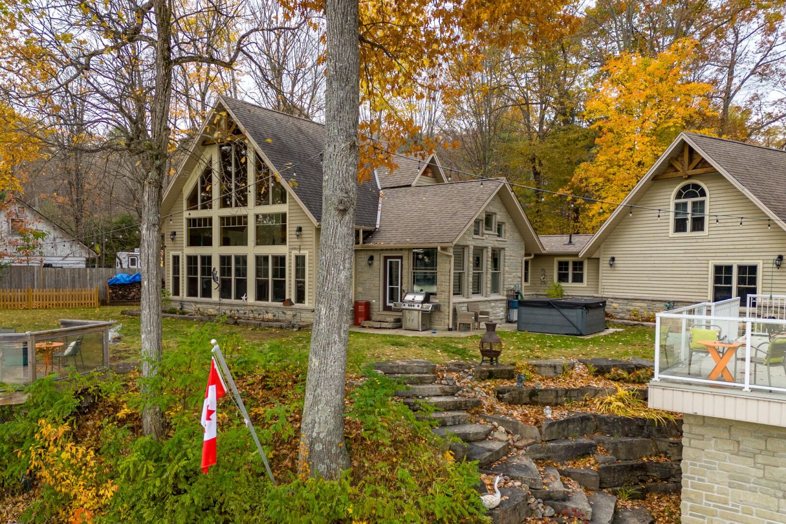 A large cottage with tons of windows sits lakeside on a green lot. Stone steps to the side lead down to a big boathouse, which has a rooftop patio at ground level.