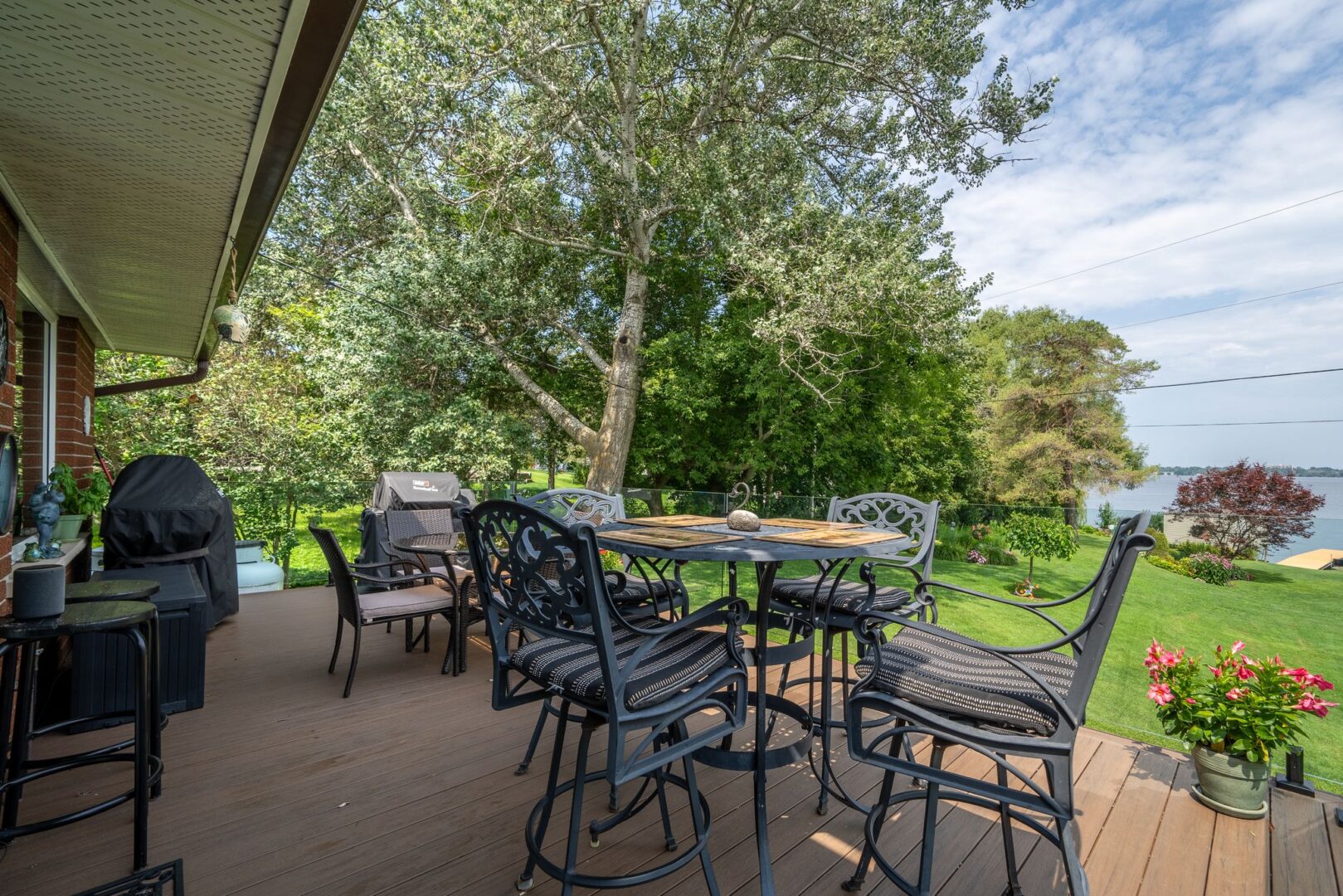 A big deck off the back of a bungalow, with a table, chairs, a glass railing, and a view of the water.