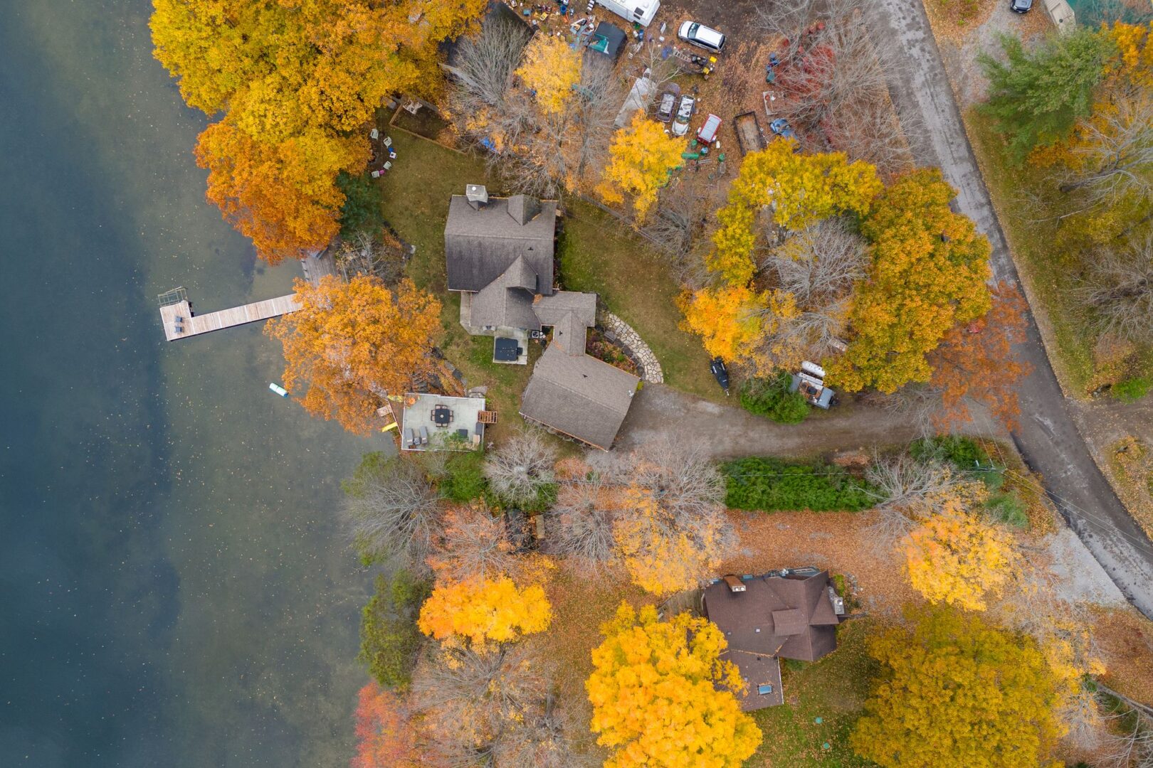Aerial view of a big cottage with a long dock that extends into a calm lake. Trees with colourful fall leaves surround the property.