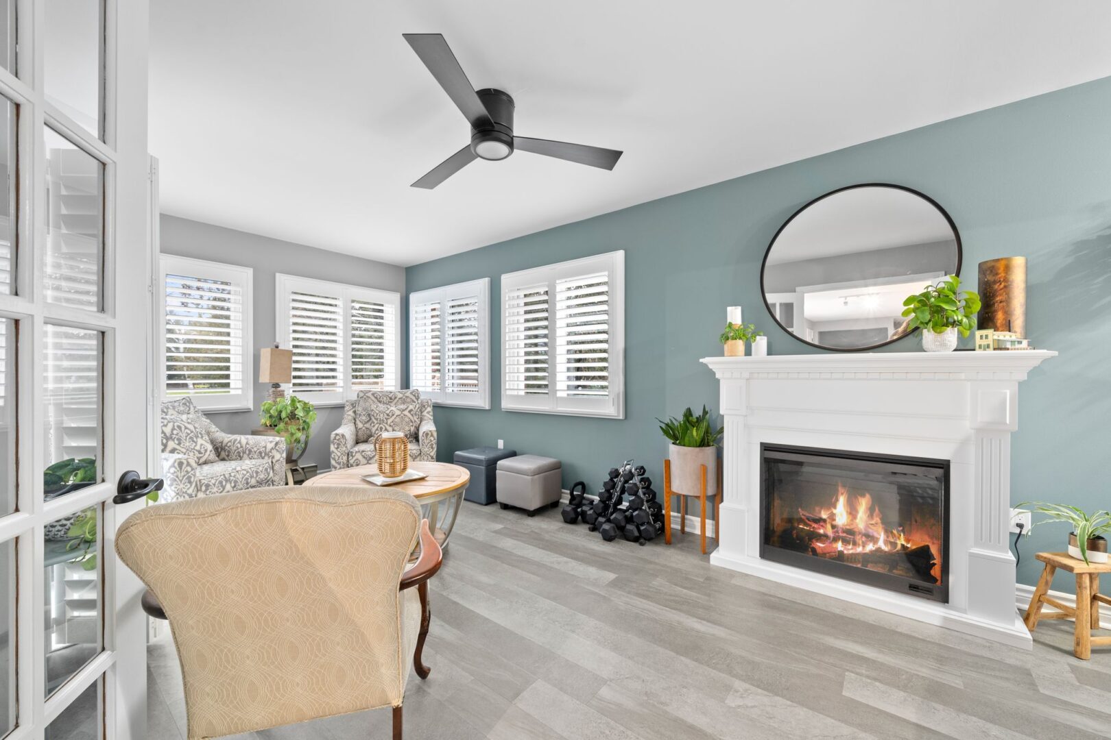 A white-painted fireplace sits in the middle of a blue wall, in a sitting area with grey vinyl plank flooring. A large round mirror hangs over the fireplace.