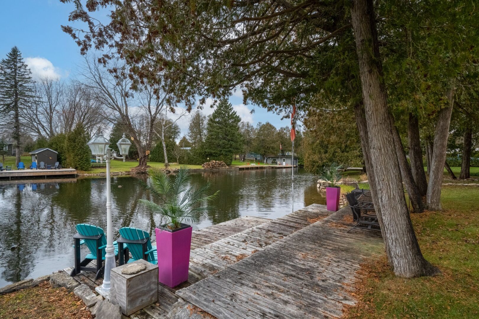 A tiered wooden deck with three levels leads from a grassy yard down to a river. Pink planters and blue Muskoka chairs sit on the deck.