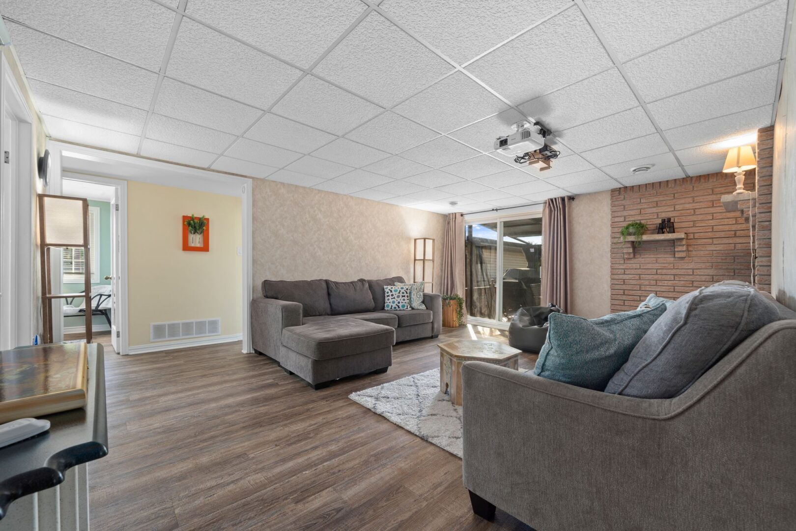 A wide living area with grey couches, wood-panelled flooring, double doors leading out to a deck, and a projector mounted to the ceiling.