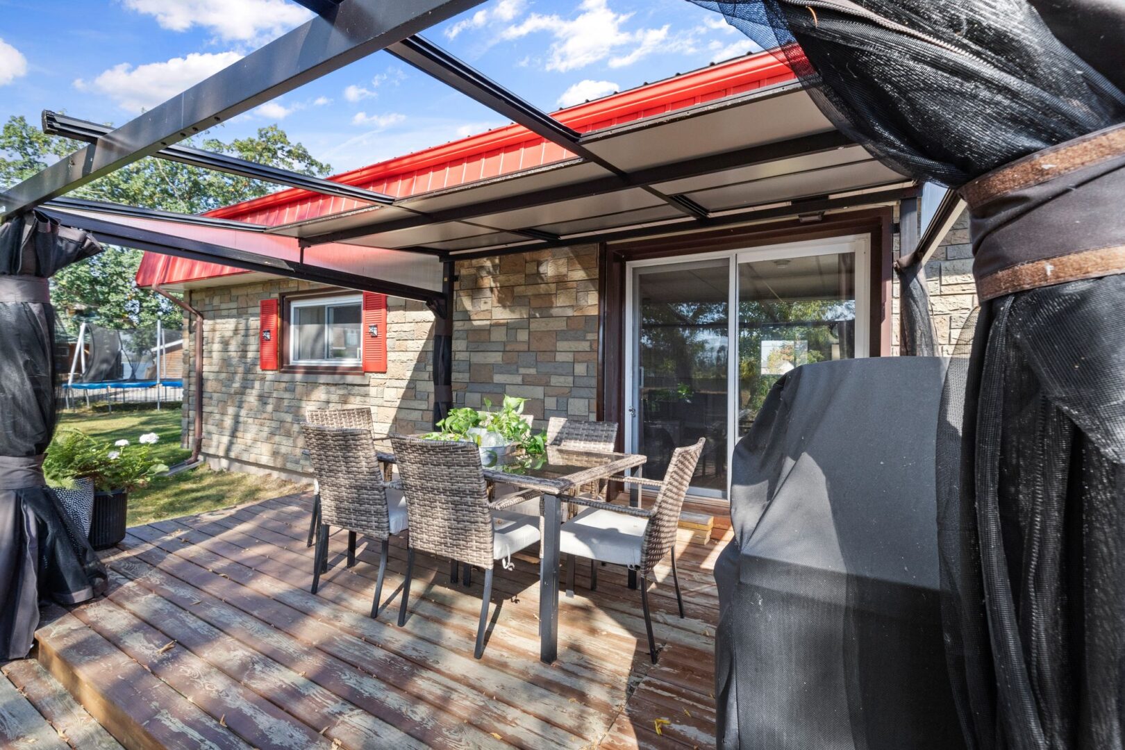 A covered deck extends off the side of a bungalow. There is outdoor dining furniture and a barbecue on the deck.