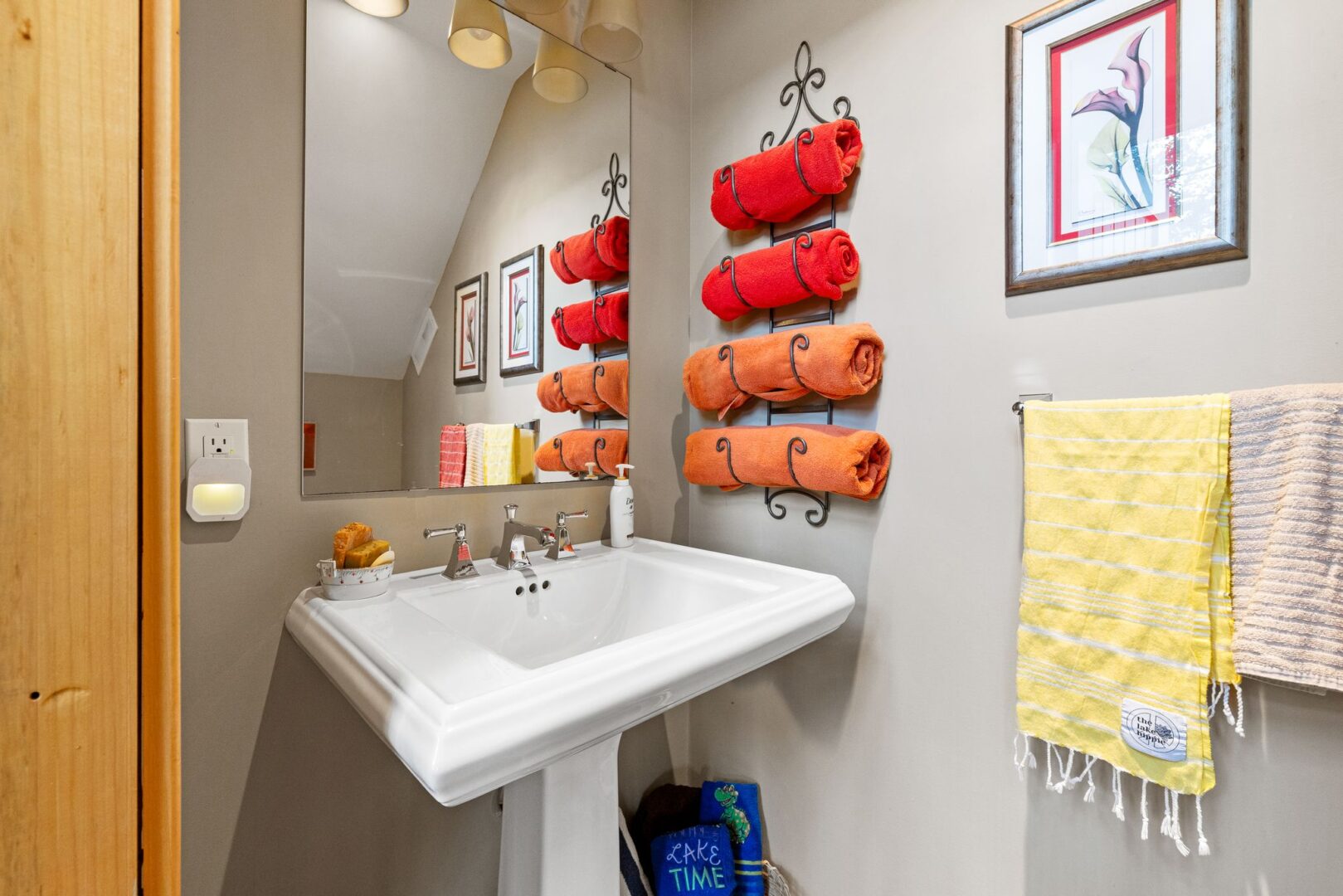 A small bathroom with with a sink. Red and orange towels hang rolled up on the wall across small hooks.