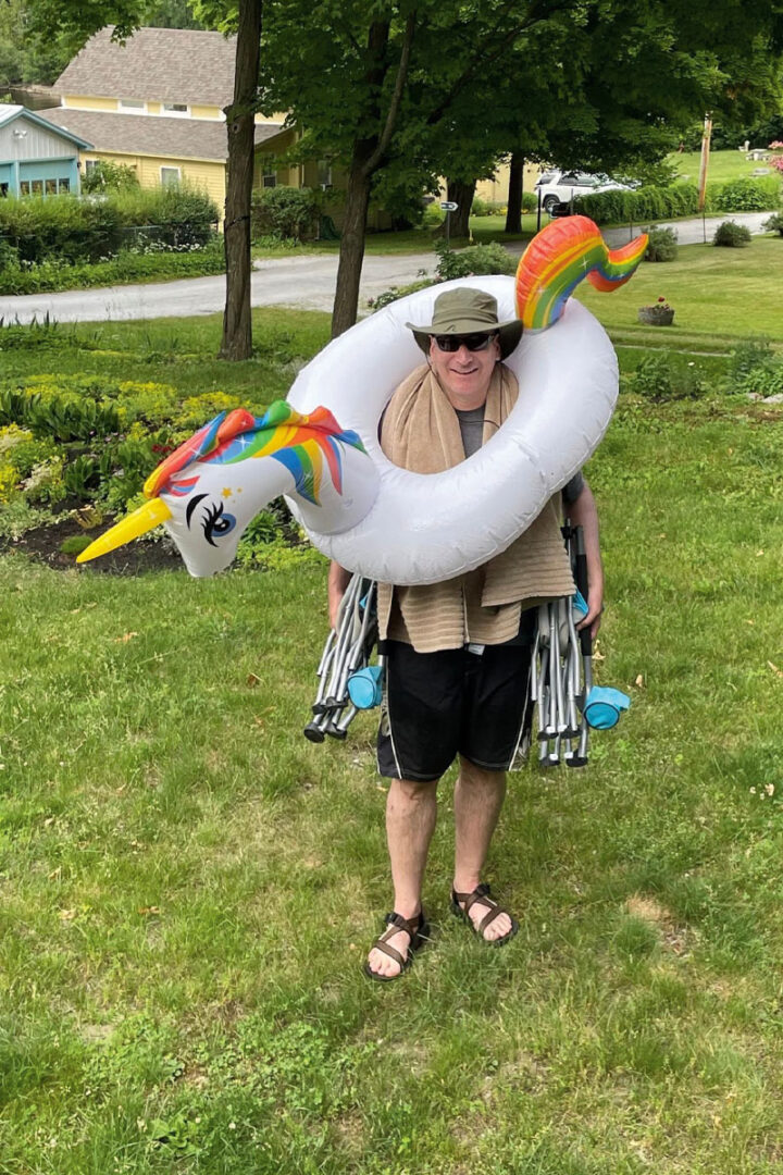 A man standing on land carrying two folded-up camp chairs and wearing an inflatable unicorn pool toy around his chest.