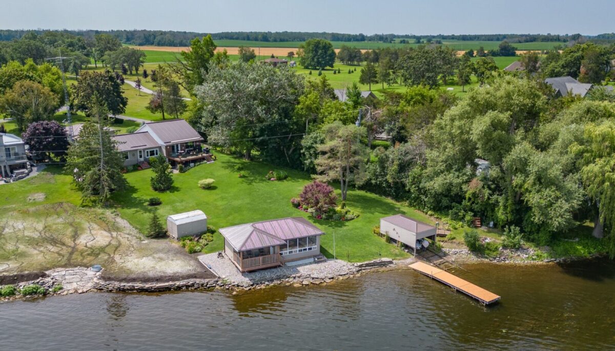 A residential waterfront property. A bungalow sits at the back of a sloping green space. By the shoreline there is a bunkie, a dock, and a small boat launch with a rail system.