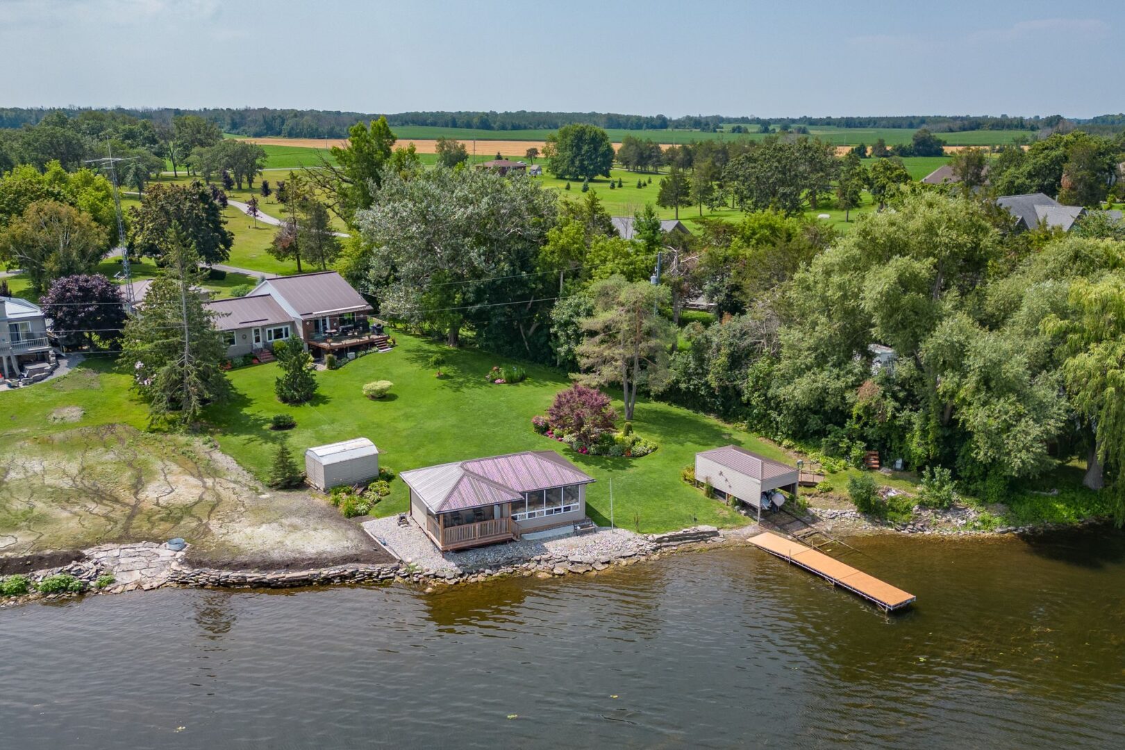 A residential waterfront property. A bungalow sits at the back of a sloping green space. By the shoreline there is a bunkie, a dock, and a small boat launch with a rail system.