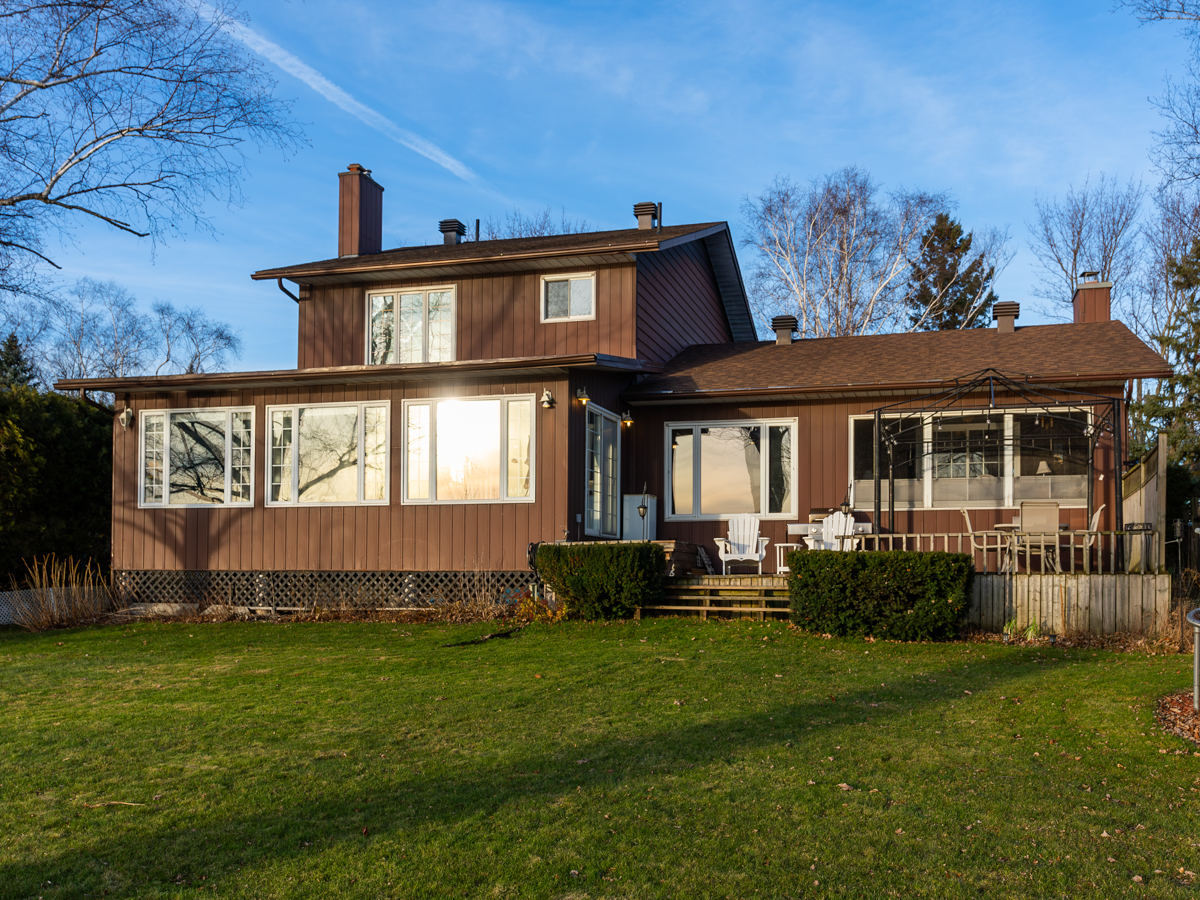 A two-story home with a brown exterior and lots of windows sits on a lakeside grassy lot.