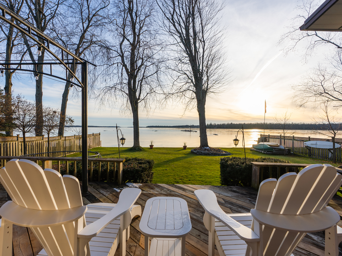View from two white Muskoka chairs on a raised deck, looking out across a big grassy area and a lake beyond.