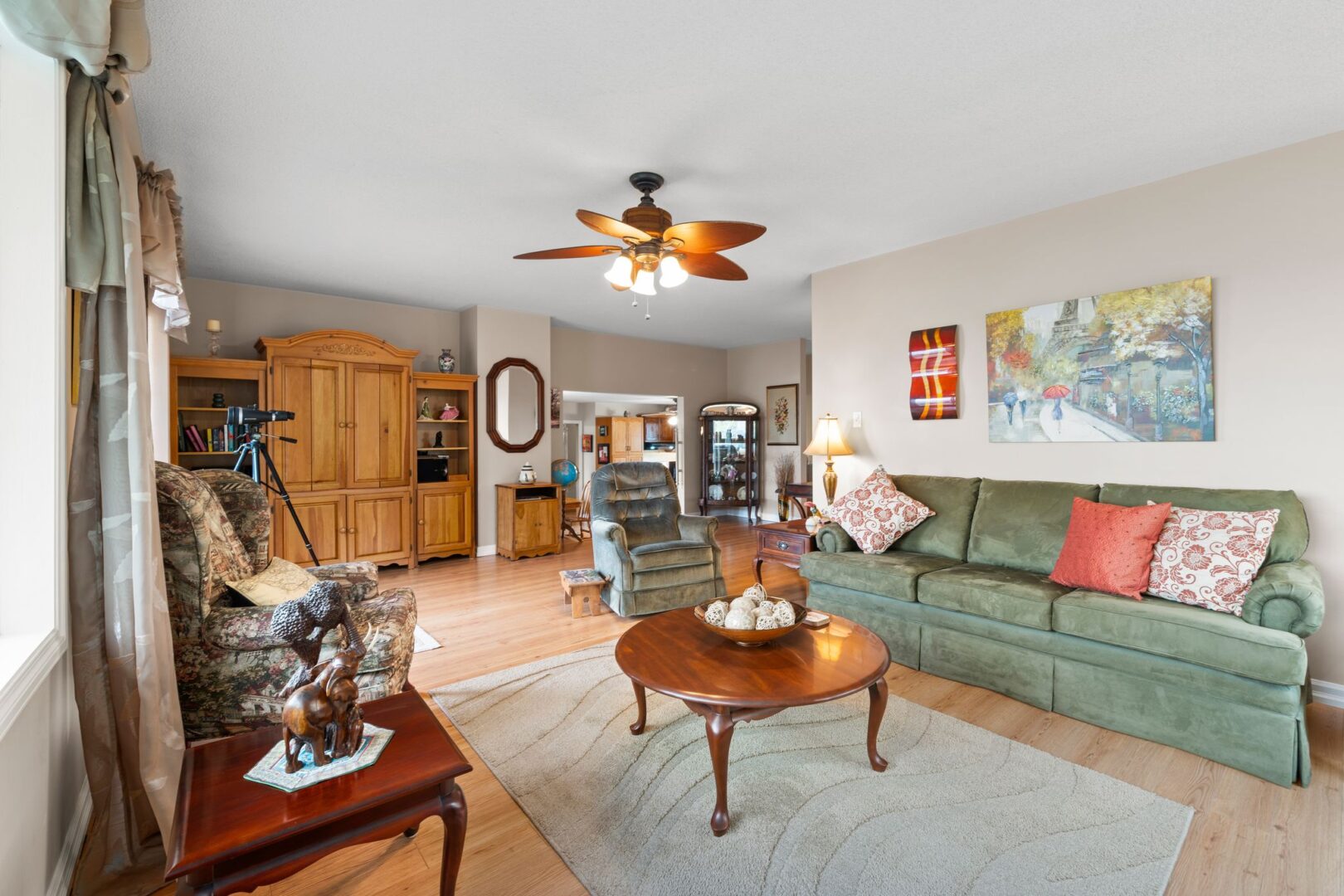 A bright living area with hardwood flooring, a couch, two chairs, a ceiling fan, and a big window.