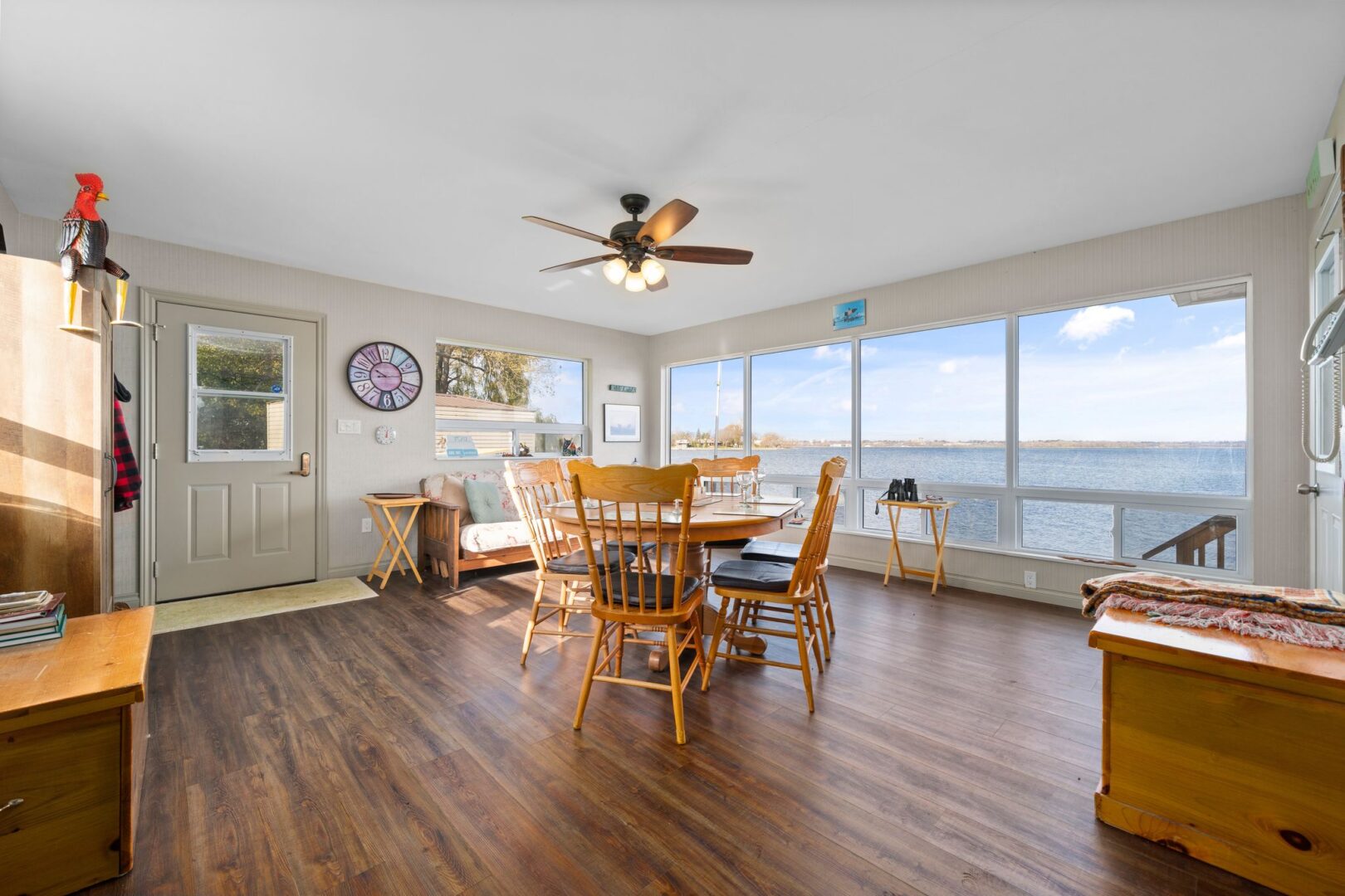 A bright interior space of a waterfront bunkie, with wood flooring, a table and chairs, a couch, a ceiling fan, and a big window facing the water.