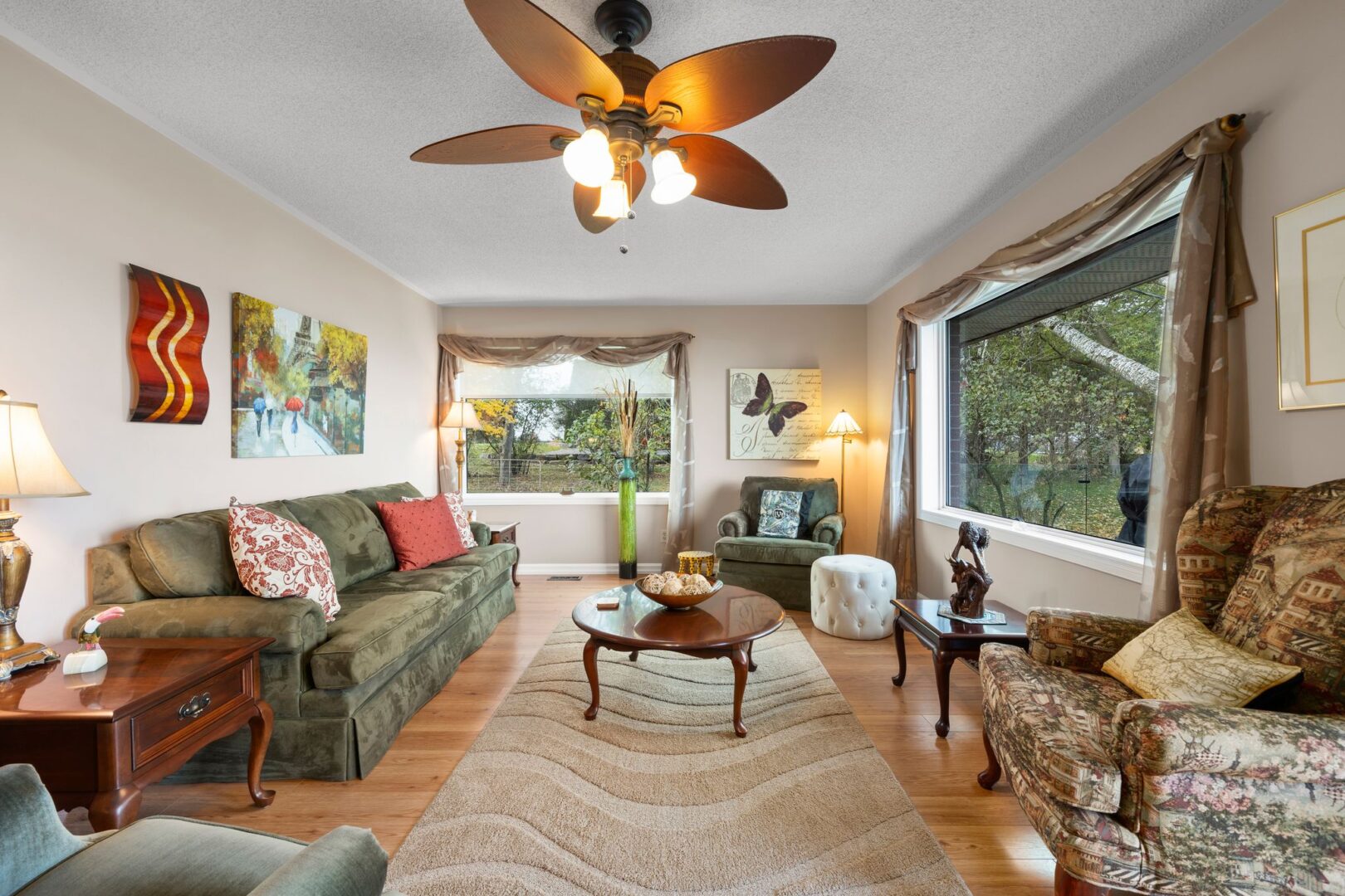 A bright living area with hardwood flooring, a couch, two chairs, a ceiling fan, and a big window.