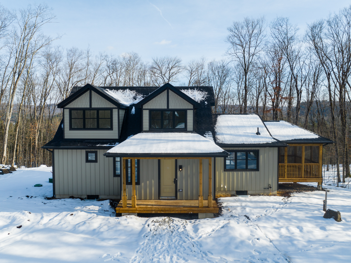 Front of a large two-story house in a snowy, rural area. The house has big windows with black trim and a wood front porch.