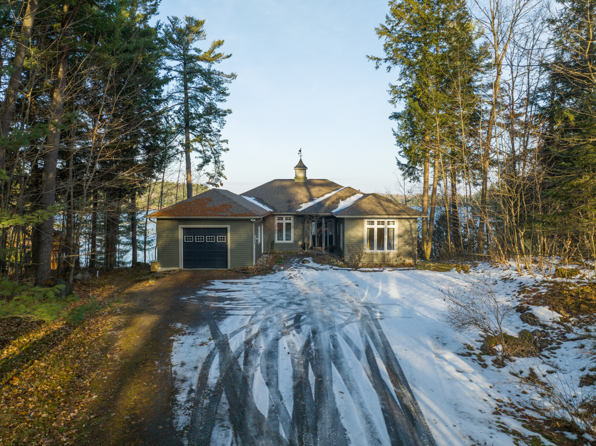 A large home with a big driveway sits in between tall trees. There is snow on the ground.