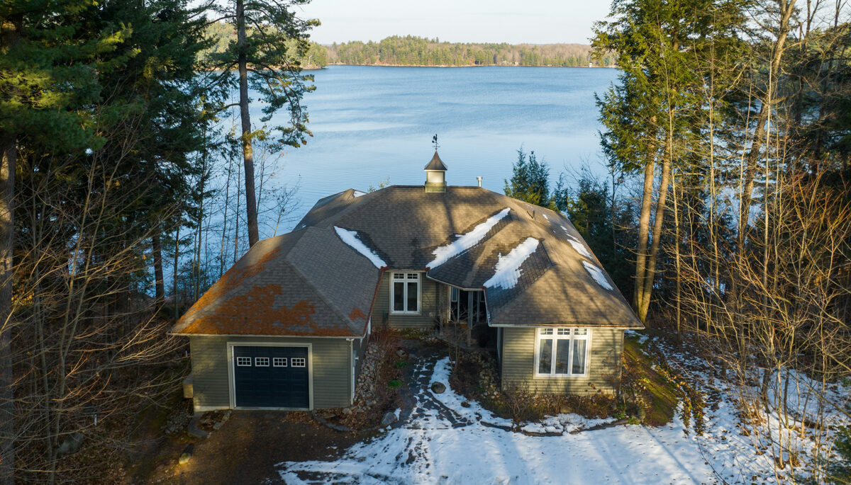 A large home with a big driveway sits at the edge of a lake, in between tall trees. There is snow on the ground.