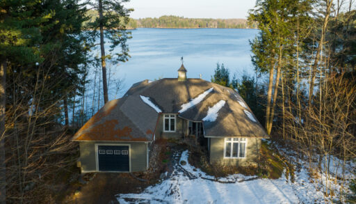 A large home with a big driveway sits at the edge of a lake, in between tall trees. There is snow on the ground.