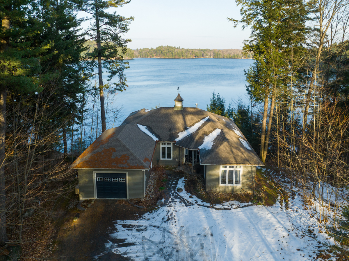 A large home with a big driveway sits at the edge of a lake, in between tall trees. There is snow on the ground.