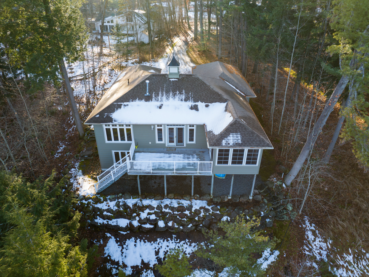 Overhead view of a large home with lots of windows that sits at the edge of a lake, in between tall trees. There is snow on the ground.