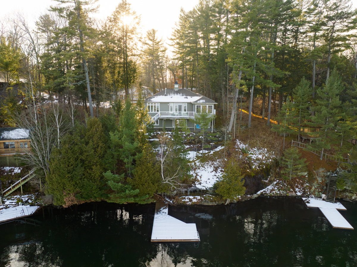 A large cottage sits waterfront on the shore of a lake. Green trees surround it on either side. A big snow-covered dock extends into the water.