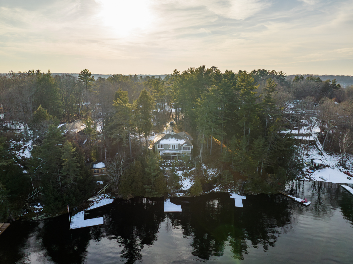 Waterfront of a lake lined with green trees and large homes. Big docks extend into the water along the shore, and there is snow covering them.